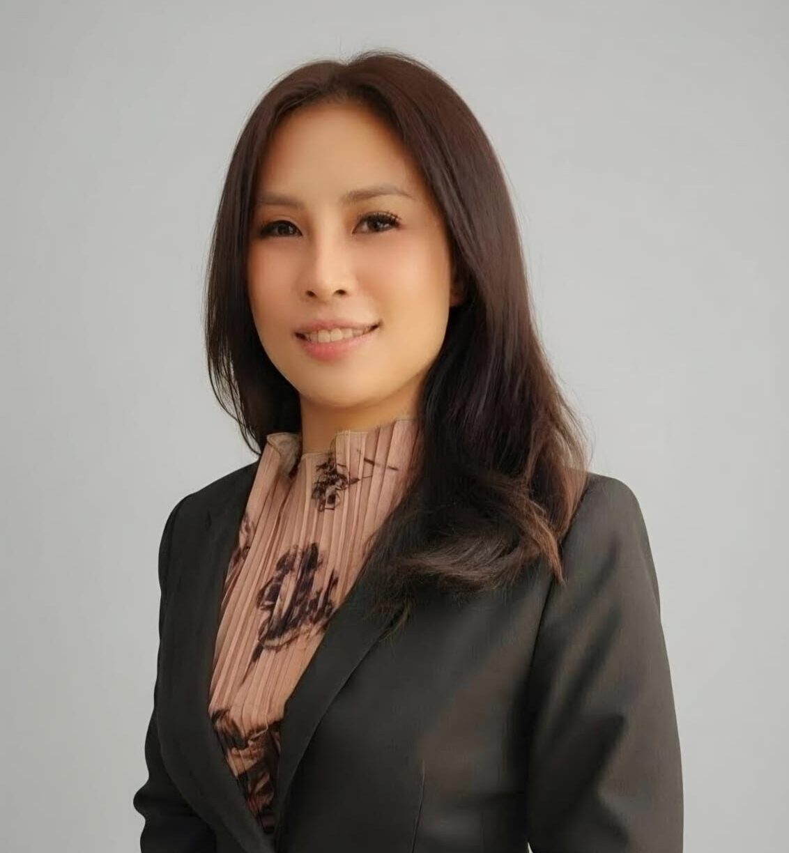 Professional portrait of a woman with shoulder-length dark hair, wearing a black blazer over a pink patterned blouse, smiling against a light gray background.