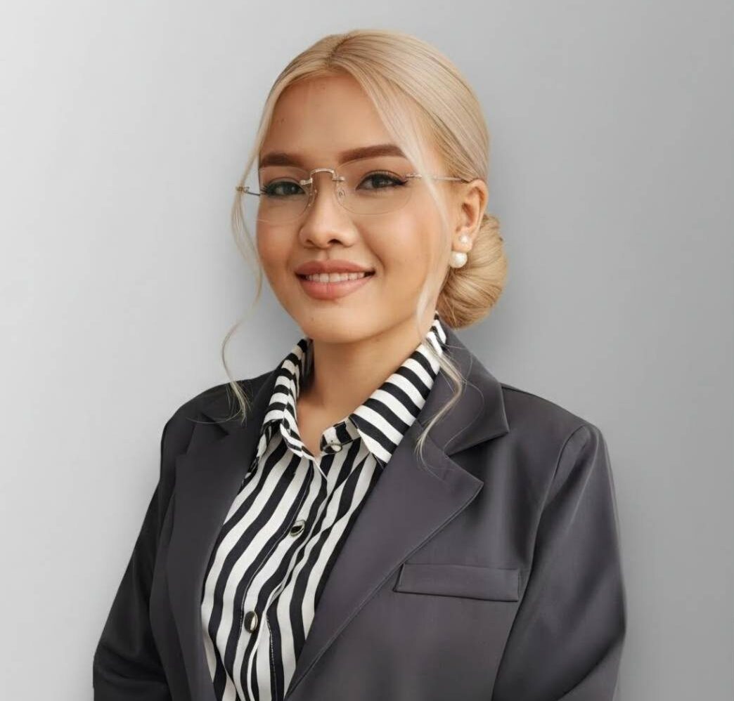 Professional woman with blonde hair in a low bun, wearing glasses, a striped blouse, and a dark blazer, smiling against a gray background.