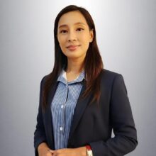 Professional headshot of a woman in a navy blazer and blue striped shirt, standing against a gray background, with hands in front.