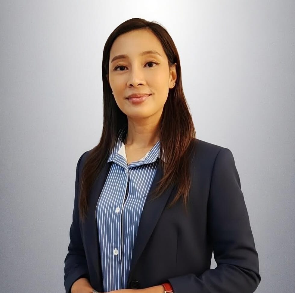 Professional headshot of a woman in a navy blazer and blue striped shirt, standing against a gray background, with hands in front.