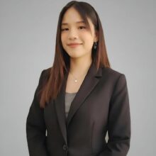 Professional portrait of a young woman in a black blazer, smiling softly against a neutral gray background.