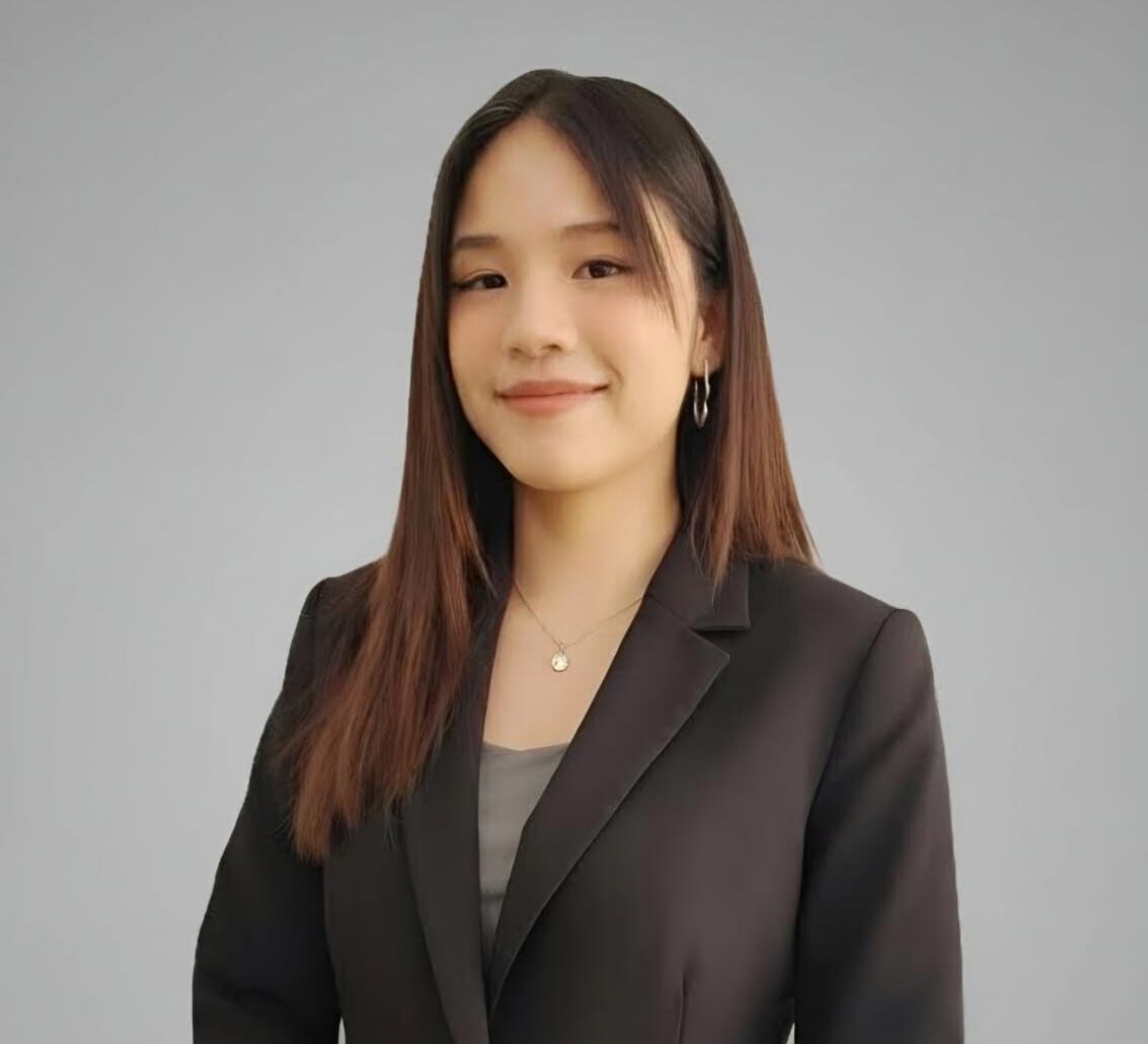 Professional portrait of a young woman in a black blazer, smiling softly against a neutral gray background.