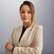 Professional woman with arms crossed, wearing a beige blazer and black blouse, posing against a gray background.