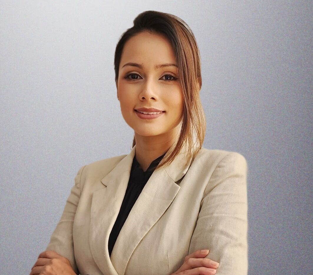 Professional woman with arms crossed, wearing a beige blazer and black blouse, posing against a gray background.